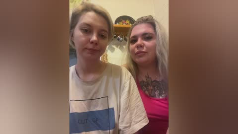 Video of two women standing side by side, one with short blonde hair in a white T-shirt, the other with long blonde hair in a pink top, both smiling, in a home setting with a wooden shelf visible in the background.