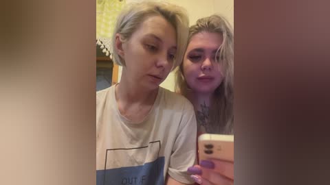 Video of two women with short hair, one in a white T-shirt, the other with long blonde hair, standing closely, taking a selfie in a bathroom with a mirror.