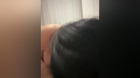 Video of a person with black hair leaning forward, viewed from behind, partially obscured by two vertical panels. The background features white vertical blinds.