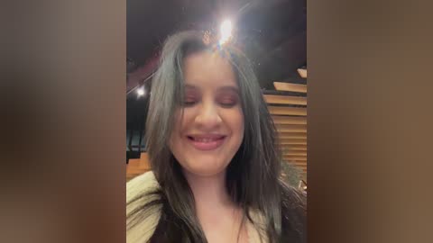 Video of a young woman with long, wavy black hair, wearing a green top, smiling and closing her eyes in a cozy room with wooden walls and ceiling beams.