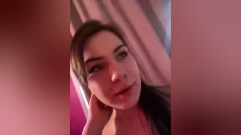 A video of a young woman with fair skin, dark hair, and dark eyes, looking contemplative, taken from a close-up angle, with blurred figures and pink and purple hues in the background.