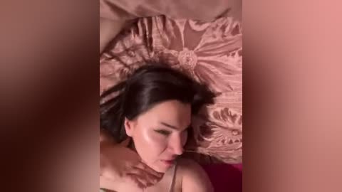 Video of a woman with dark hair lying on a peach-colored satin bedspread, eyes closed, wearing a brown bra.