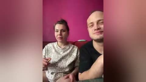 A video of a smiling, bald Caucasian man in a black t-shirt and a Caucasian woman with dark hair in a white polka-dot shirt sitting side by side against a pink wall.