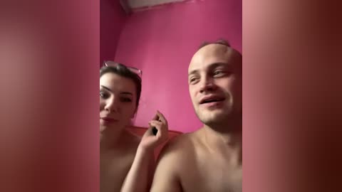 Video of a topless, bald white man and a topless woman with brown hair, both smiling, in a pink room with mirrored walls.