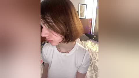 Video of a young woman with straight, shoulder-length brown hair, wearing a white t-shirt, in a dimly lit bedroom with beige walls, a bed, and a framed picture on the wall.
