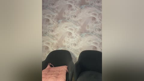 Video of a person\u2019s feet in black shoes with a pink blanket draped over them, standing on a textured, swirling, white and light pink marble-like floor.