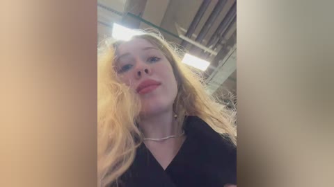 A video of a young woman with long, wavy blonde hair and pale skin, wearing a black top, standing in a dimly lit room with industrial ceiling beams.