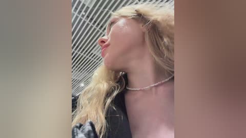 Video of a blonde woman with wavy hair, fair skin, and a thin body, seen from the side. She wears a pearl necklace and has a pouty expression. The background features a grid-patterned ceiling and blurred objects.