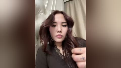 A video of a young Asian woman with shoulder-length, wavy, dark brown hair, wearing a black lace-trimmed top. She has a serious expression, and her hand is raised in front of her face, possibly to adjust her hair. The background features beige curtains and a white wall.