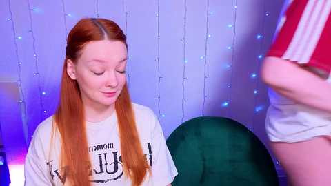 Video of a fair-skinned redhead girl in a white T-shirt, sitting on a green chair, facing a man in red and white striped shorts, with blue string lights in the background.