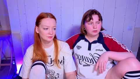 Video of two young women with fair skin, red hair, and medium builds, wearing white sports jerseys. One sits, the other stands behind, in a dimly lit room with string lights and a blue light source.