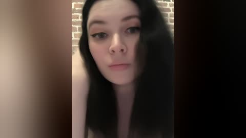 A video of a young, pale-skinned woman with long, dark hair, wearing minimal makeup, looking directly into the camera against a brick wall background.