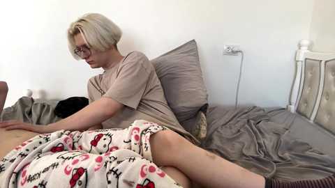 Video of a pale-skinned, short-haired woman in glasses wearing a beige t-shirt and red pajama shorts, lying on a messy gray bed with pillows and a white blanket with cartoon characters.