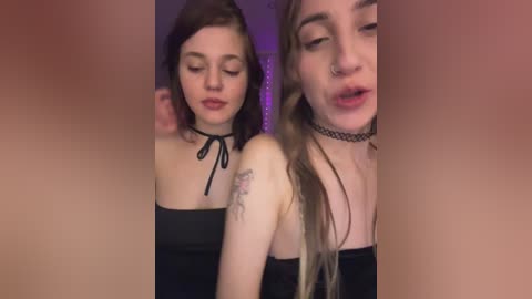 Video of two young women with fair skin and medium-length hair, one with a choker and the other with a black top, in a dimly lit room with a purple backdrop.