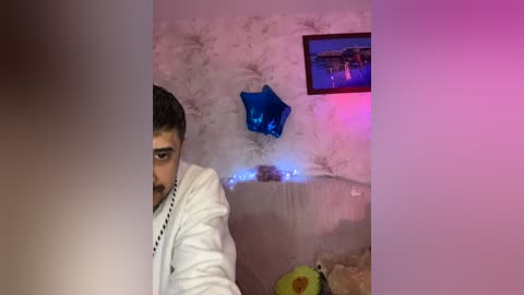 Video of a man with dark hair, beard, and glasses, wearing a white jacket, standing in a dimly lit room with pink and purple lights, a blue star decoration, and a framed picture on the wall.