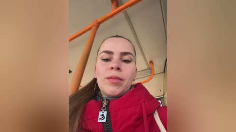 Video of a woman with light skin and brown hair, wearing a red jacket, standing inside an orange subway car.