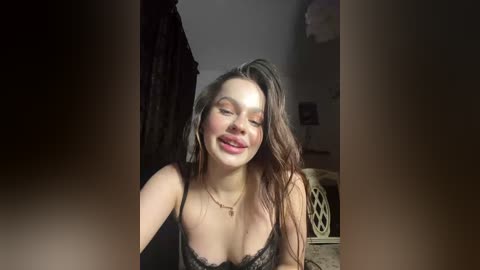A video of a young woman with long brown hair, wearing black lace lingerie, smiling with her eyes closed, in a dimly lit room with a bed and a patterned wall.
