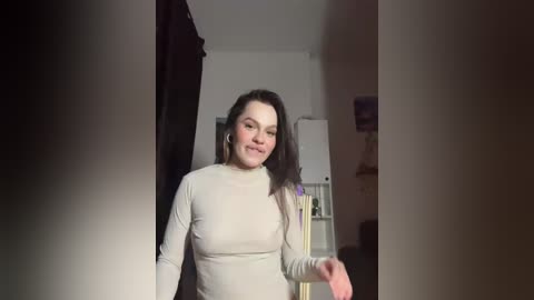 Video of a smiling woman with medium-length black hair, wearing a beige turtleneck, standing in a dimly-lit hallway.