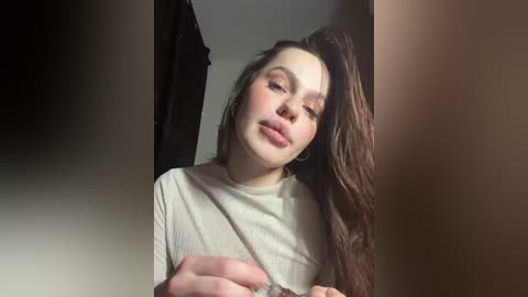 Video of a young woman with long, wavy brown hair, wearing a beige shirt, gazing downwards, in a dimly lit room with a dark curtain in the background.