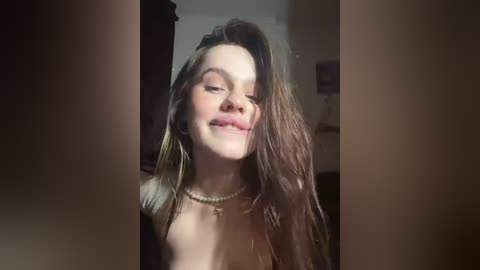 A video of a smiling, fair-skinned woman with long, wavy brown hair, wearing a pearl necklace, standing in a dimly lit room.