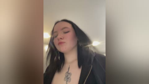 Video of a fair-skinned, dark-haired young woman with shoulder-length hair, wearing a black jacket, and a prominent \"Fuck You\" tattoo on her chest, standing indoors with a blurred background.