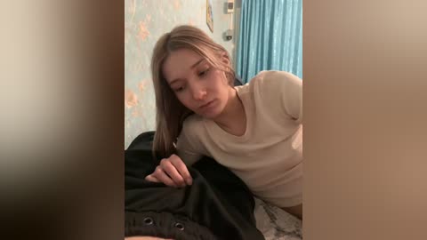 A video shows a young woman with light skin and long, straight, blonde hair, wearing a beige long-sleeved shirt, lying on a bed with black and gray blankets. The room has pastel floral wallpaper and a blue curtain.