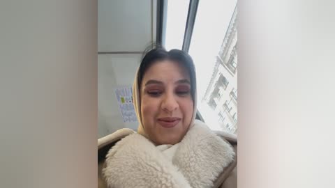 Video of a woman with light skin, dark hair, wearing pink eyeshadow, and a beige scarf indoors near a window, showing a blurry urban street outside.