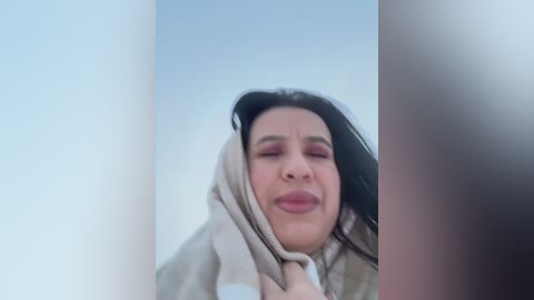 Video of a woman with closed eyes, wearing a beige hooded sweatshirt, against a blurred, light-colored background. Her expression appears pensive or contemplative.