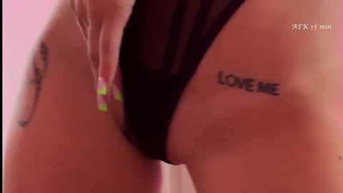 Video of a woman's lower body, wearing black sheer lingerie. Her left hand, with neon green nails, is pulling down the lingerie, revealing her pubic area. \"LOVE ME\" tattoo on her left hip.