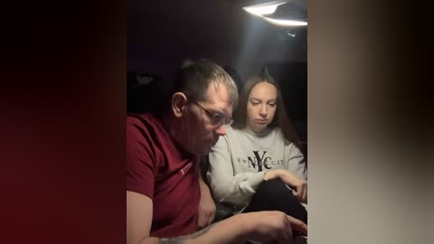 Video of a middle-aged man with glasses and a maroon shirt, and a young girl with long hair, both intensely focused on a tablet, in a dimly lit room with a ceiling light.
