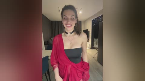 Video of a smiling Asian woman with straight black hair, wearing a black strapless top, red draped fabric, black choker, in a modern, dimly-lit living room with dark furniture.