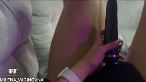 Video of a woman with light skin and medium build, wearing a white wristband, using a black vibrator on her thigh. Text overlay reads \"BBR\" and \"MILLY ENA VAGINONA.\