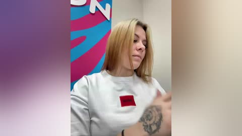 A video of a blonde woman with straight hair, wearing a white sweatshirt with a red square emblem, and a rose tattoo on her wrist, in a brightly colored, abstract background.