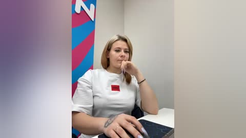 Video of a young Caucasian woman with shoulder-length blonde hair, wearing a white T-shirt with a red logo, sitting in a sterile, beige-walled room.