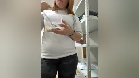 Video of a fair-skinned woman with shoulder-length auburn hair, wearing a white shirt and black pants, holding a clear plastic container with a yellowish substance, standing in front of a white shelving unit filled with assorted items in a cluttered room.