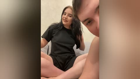 A video of a young woman with long black hair, wearing a black shirt and shorts, sitting cross-legged on a gray couch in a dimly lit room.