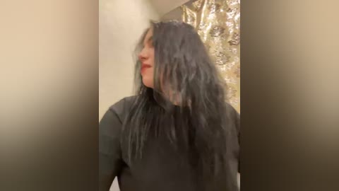 Video of a woman with long, wavy black hair and red lipstick, standing against a beige wall with a gold textured background. She wears a black top.
