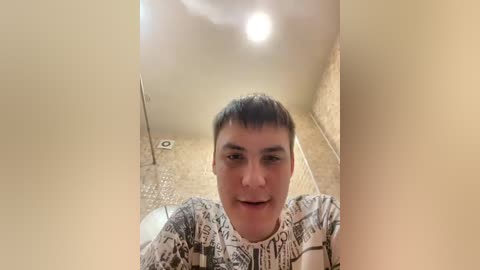 Video of a young man with short dark hair, wearing a comic book-patterned T-shirt, standing in a narrow, beige-walled hallway with a circular ceiling light, textured floor tiles, and a trash can in the background.