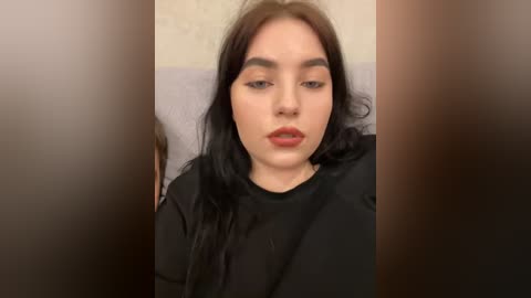 A video of a young woman with fair skin, long black hair, and full lips, wearing a black top, sitting on a light-colored couch with a blurred background.