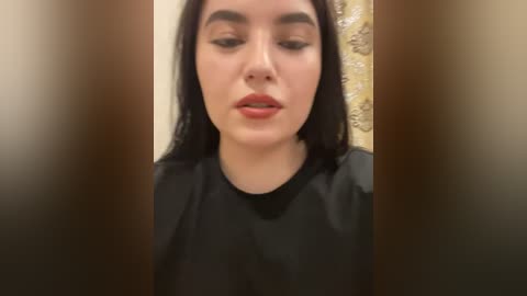 Video of a young woman with fair skin, straight black hair, and dark eyebrows, wearing a black dress, looking down, in a blurred, neutral background.