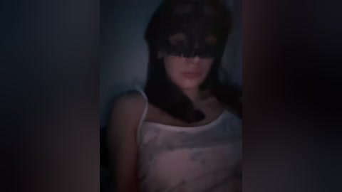 A dimly-lit video of a woman with long black hair, wearing a sheer white blouse, black lace mask, and a sultry expression, set against a shadowy background.