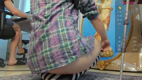 Video of a person, possibly male, wearing a plaid shirt and striped socks, kneeling on a carpeted floor in an office with a large poster of a mermaid on the wall.