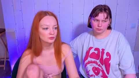 Video of two young people with fair skin. The redhead in a pink top sits, while the boy with short brown hair wears a gray \"L.A. Guns\" shirt. Background features white wall with string lights and a wooden table.