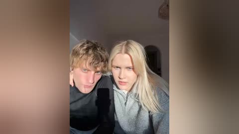 A video of a young blonde woman and a man with short brown hair, both with pale skin, wearing casual dark clothing, standing close in a dimly lit room with blurred background.