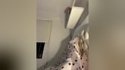 Video of a cozy, dimly lit bedroom with a white bed covered in star-patterned bedding. A small white shelf with a decorative item hangs on the wall above the bed.