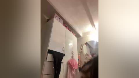 Video of a cluttered, beige-toned closet with hanging clothes, a white cabinet, and a partially visible mirror reflecting a person in a pink shirt.