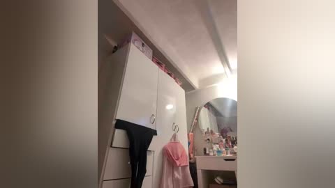 Video of a tidy, modern bathroom with white cabinets, a black towel hanging, and a pink robe on a hook. Sink area with toiletries and a mirror.
