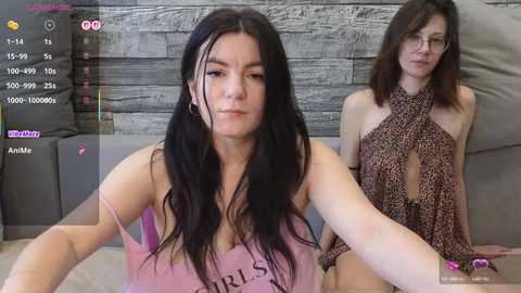 A video of two women, one with long black hair in a pink tank top, the other with glasses and a leopard-print scarf, sitting on a grey couch.