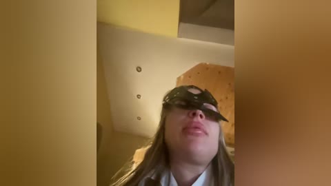 Video of a young woman with long brown hair, wearing a black mask with eyeholes, looking upwards in a dimly lit room with beige walls and a wooden ceiling.