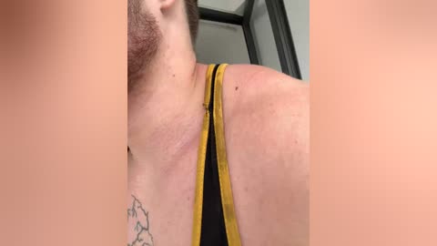 A close-up video of a light-skinned man with a beard, wearing a black tank top with yellow trim, showing a small tattoo and redness on his neck, in a blurred indoor setting.